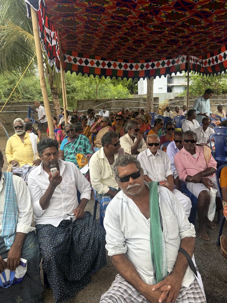 Elderly patients in sunglasses seated in blue chairs under a canopy at a community eye camp